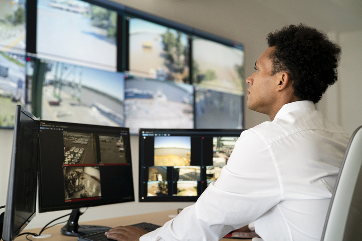 Medium shot of young adult male security worker watching video wall while sitting at desk in control room