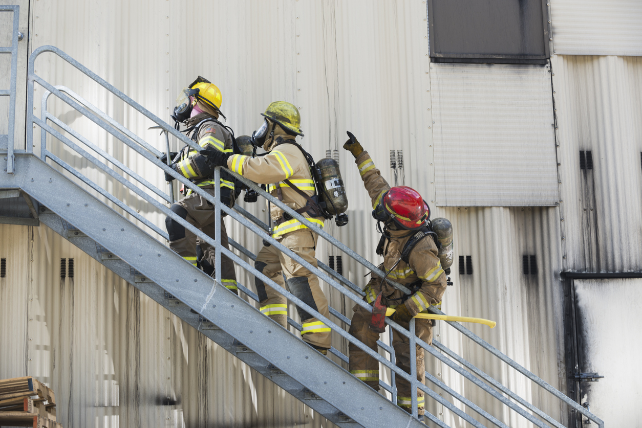 A group of three multiracial firefighters, led by a woman, climbing up a metal staircase on the exterior of an industrial building or warehouse. She Is holding a crowbar, and has a patch of an American flag on her arm. They are wearing protective clothing, helmets and oxygen masks.